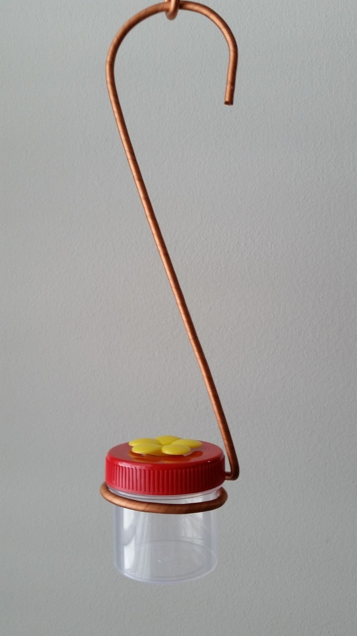 Handmade copper wire hummingbird feeder with a clear plastic jar red lid and a yellow flower detail hanging against a light gray wall