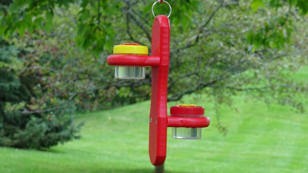 Handmade red wooden hummingbird feeder with two clear glass jars and bright yellow lids hanging outdoors against a backdrop of lush green grass and trees