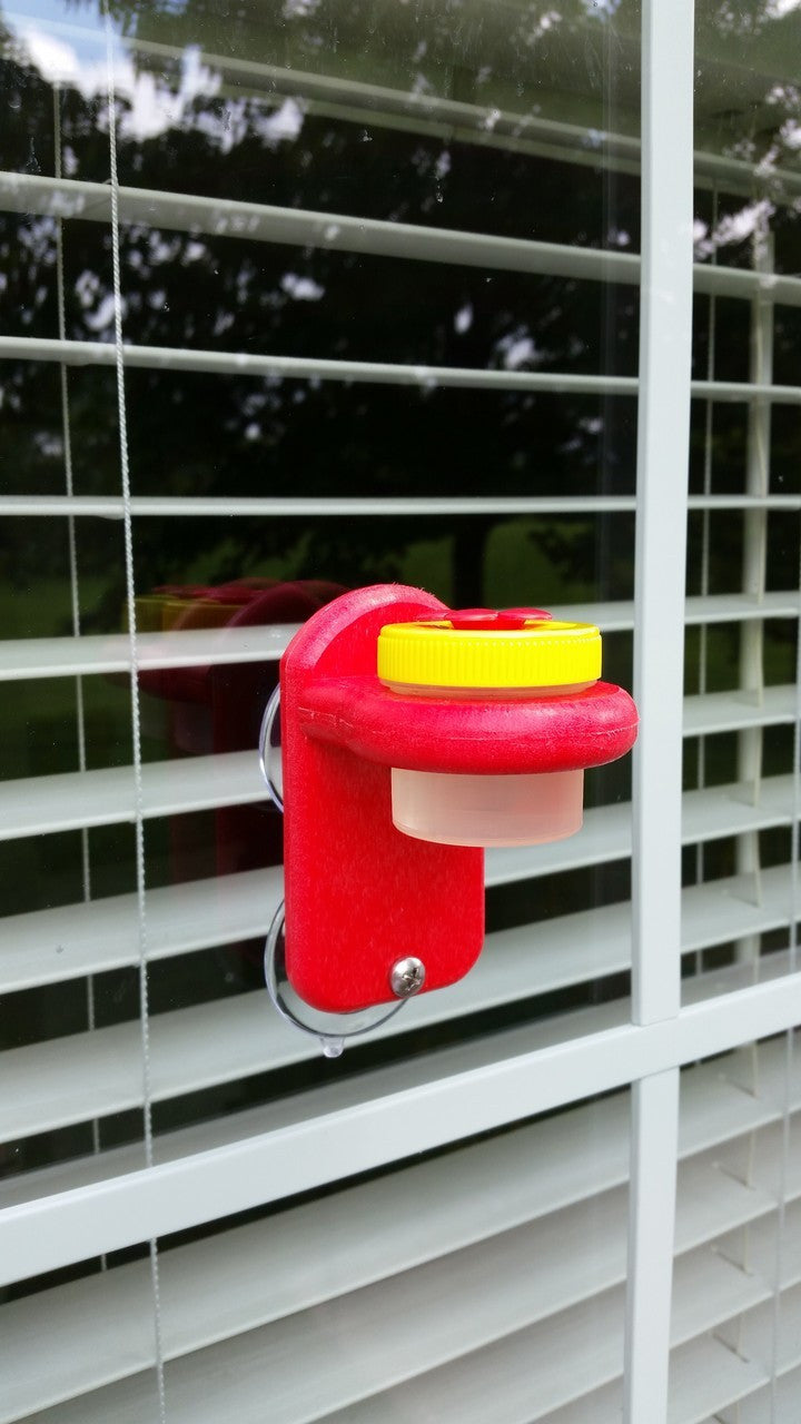 Yellow and red Nectar DOTS window hummingbird feeder attached to window with blinds, featuring large DOT holding 1.25 oz of nectar.