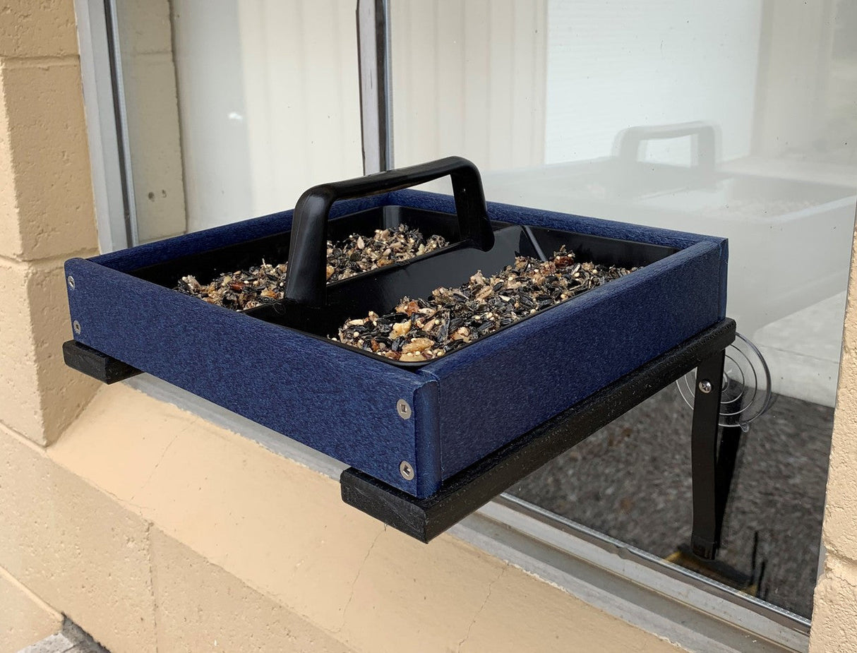 Photo of a navy blue and black recycled plastic window bird feeder with a black handle filled with birdseed affixed to a window with suction cups showcasing its divided compartments and textured surface