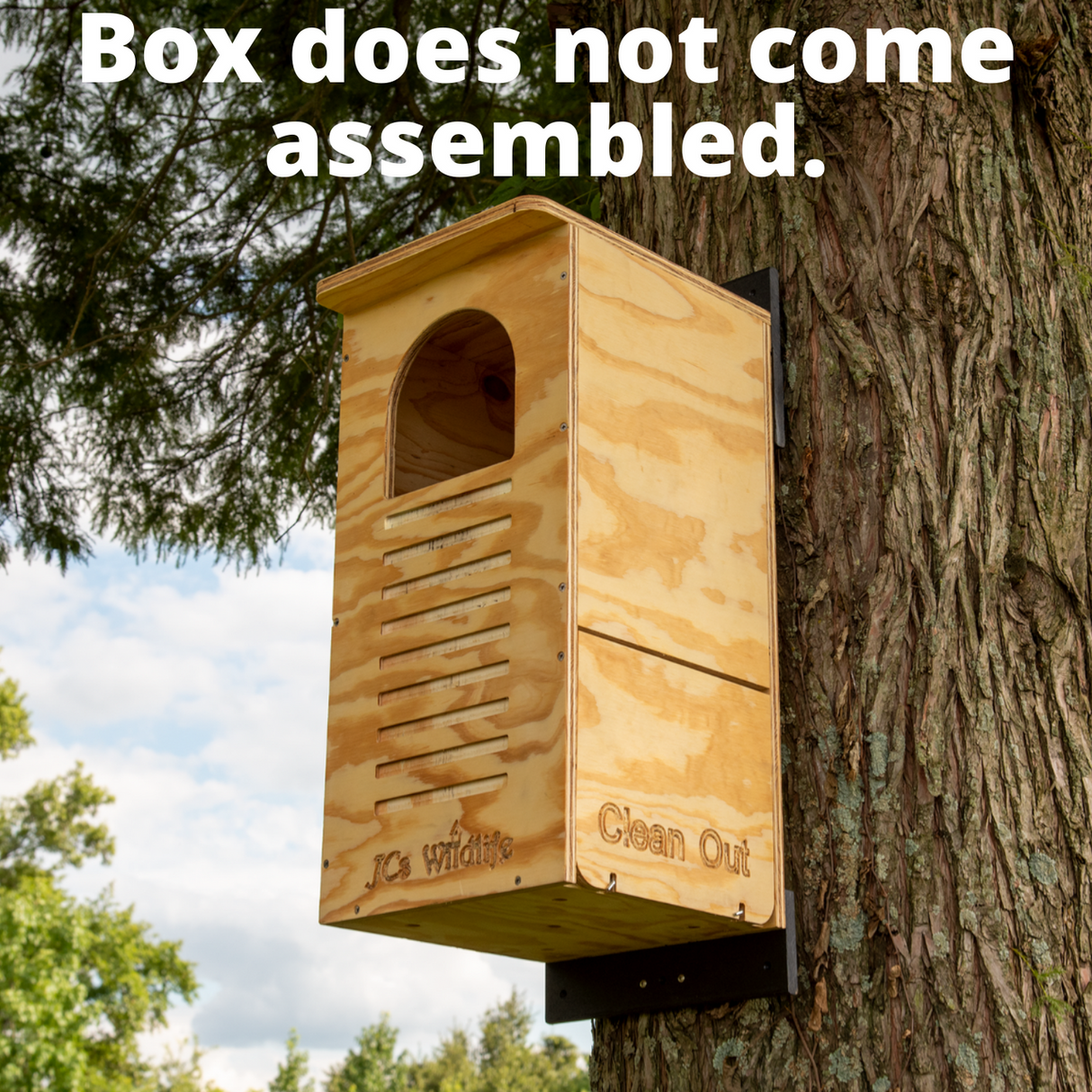 JCS Wildlife wooden birdhouse mounted on a tree outside with a clean-out door, arched entryway, and light-colored wood against a backdrop of green foliage and partly cloudy sky
