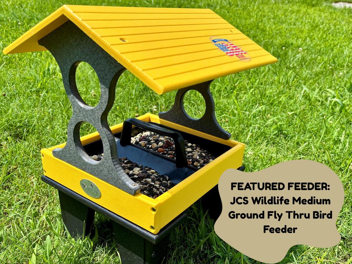 JCS Wildlife Medium Ground Fly Thru Bird Feeder photograph shows a yellow and gray plastic bird feeder on a grassy lawn featuring a USA Made emblem and filled with mixed seeds and nuts