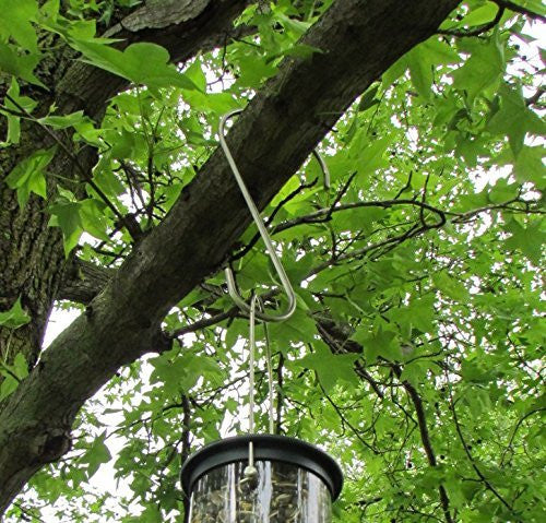 Photograph showing a stainless steel bird feeder hook hanging from a tree branch outdoors with lush green leaves and a partially visible clear bird feeder containing birdseed