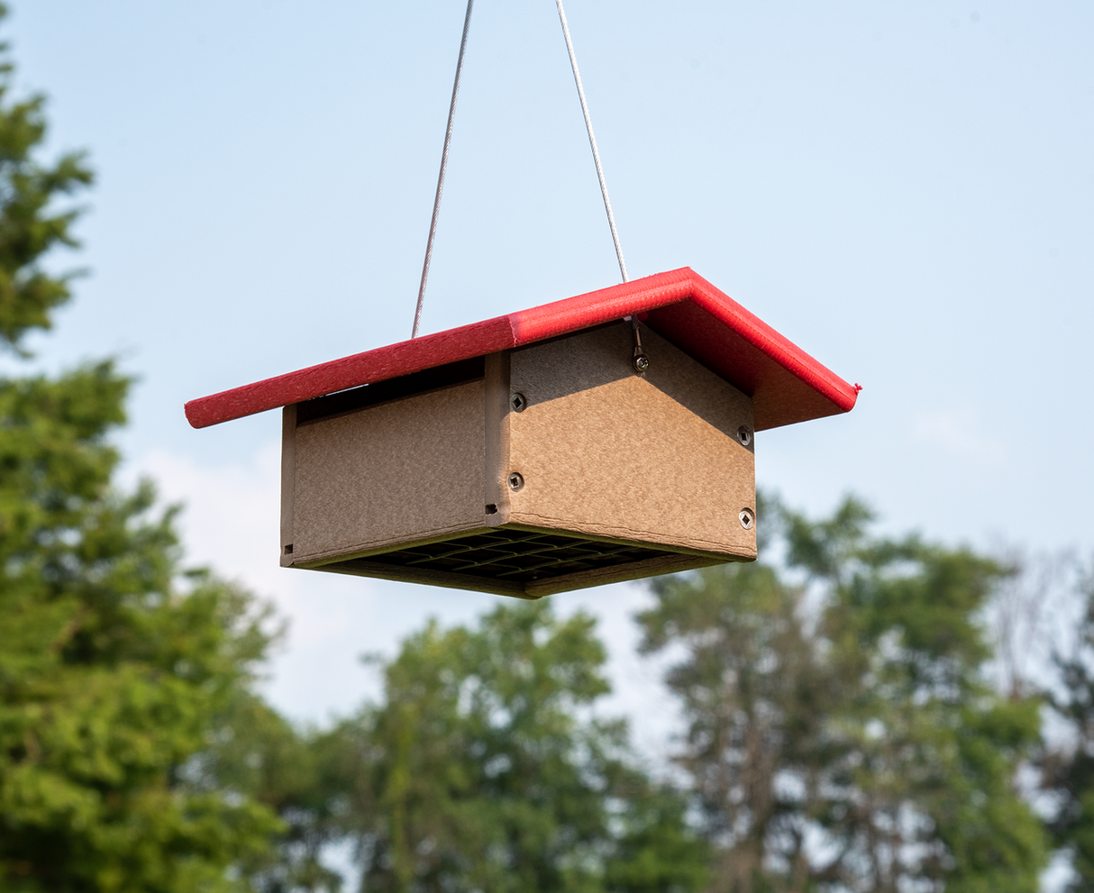 Recycled plastic bird feeder with a bright red roof hanging outdoors against a backdrop of lush green trees