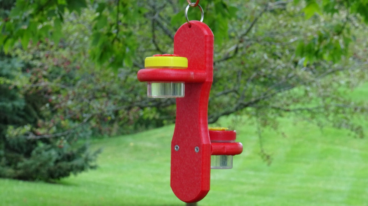 Red plastic insect trap with yellow lids hanging outdoors against a backdrop of green grass and leafy trees
