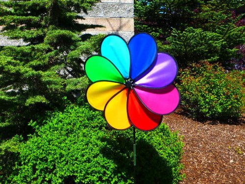 Unknown artist, vibrant rainbow pinwheel in a garden setting near lush green shrubs and evergreen trees, featuring bright red, yellow, and blue petals