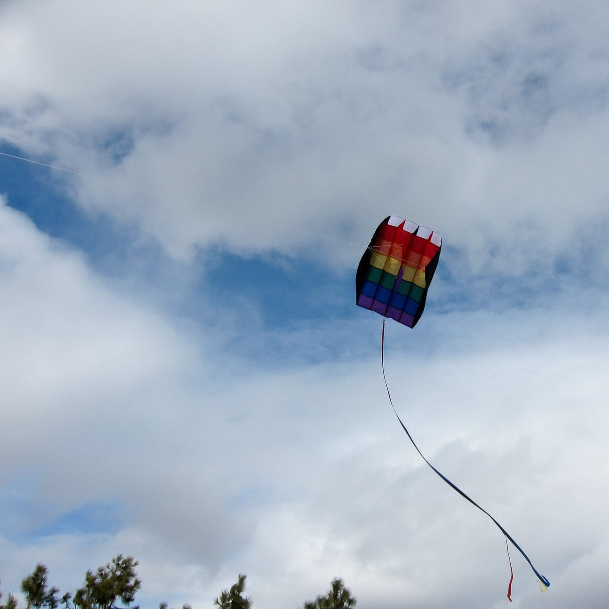 Rainbow stripes air foil kite flying in the sky with trees visible below