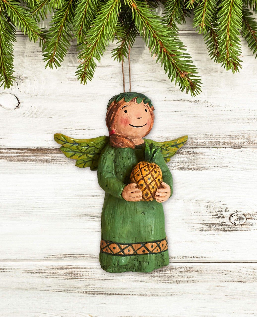 Hand-painted Studio M Warm Welcome Angel Ornament hanging from greenery, featuring a hand-carved style and holding a pineapple.